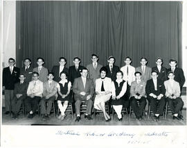 Interior, class photo, Montreal Hebrew Academy graduates, first row of students seated on chairs, second row of male students standing, a male and a female teacher seated in centre of first row. Curtain in background.