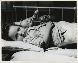Interior, young girl (approx. age 2) gazes directly at photographer, reclined on hospital bed clutching doll, lower torso and legs are encased in cast and bandages, metal bedframe in background.