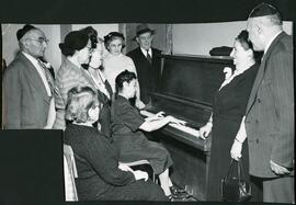 Interior group snapshot, Unidentified men and women standing and looking on at woman playing upright piano at Golden Age Centre choir rehearsal.
