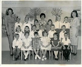 Class photo, interior, Val Royal nursery, first row of students seated on chairs, second row of students standing, third row of students standing on raised surface, a female teacher standing in back on either side. Spring decor on wall.