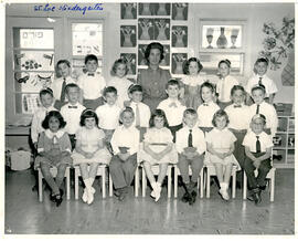 Class photo, interior, Cote St-Luc, kindergarten, first row of students seated on chairs, second row of students standing, third row of students standing on elevated surface, teacher standing in middle of third row, Purim and spring decor on wall.