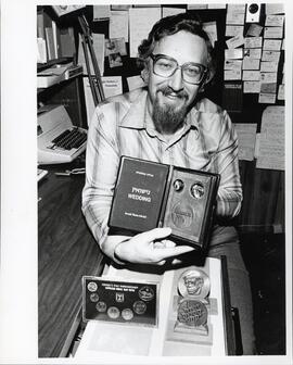 Interior portrait, Isidore Baum sitting at desk proudly displaying a wedding Israel State Medal from his large collection of Israeli coins. Israel's 31st anniversary official mint set from 1979 rests on a pull out drawer along with two other Israel State Medals.