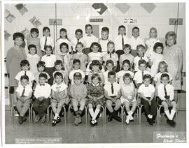 Class photo, interior, Chomedey nursery A., first row of students seated on chairs, second row of students standing, third row of students standing on a raised surface, fourth row of students standing on a higher raised surface, one female teacher standing in back on either side. Spring decor on wall.