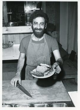 Interior portrait of Marc Fagen, former owner of L'Herberie, in kitchen wearing apron and holding plate with "smoked meat" tofu sandwich and pickle.