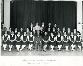 Interior, class photo, Montreal Hebrew Academy graduates, first row of female students seated on chairs, second row of male students standing, third row of male students standing on raised surface. A male and a female teacher seated in centre of first row. Curtain in background.