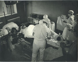 Interior, six people wearing face masks, caps and gowns, gathered in surgery room with patient reclined on hospital bed, patient wears face mask attatched to ventilation tubes that are being monitored by a person leaning over and two tanks are attached, one person stands with hands clapsed as if in prayer, hope or dread, others stand watching wearing gloves or hold towels as two individuals perform surgery upon patient.