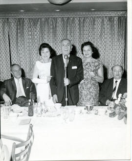 Interior group portrait of men and women seated around a set table. One man and two women standing at microphone. From the collection of the Workmen's Circle.