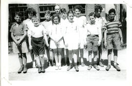 Exterior group portrait, students standing in two rows, a male teacher standing in back. Building in background.
