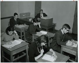 Group portrait of young adults writing test, supervisor sitting at desk in background, Jewish Vocational Services, Montreal.