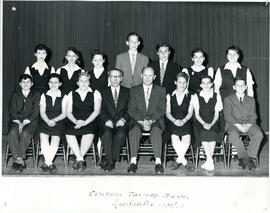 Class photo, interior, graduates, first row of students seated on chairs, second row of students standing, two male teachers seated in centre of front row. Curtain in background.