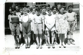 Class photo, exterior, students standing in three rows, a male teacher standing in back. A brick building in the background.
