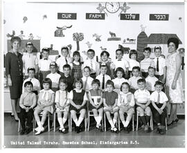 Class photo, interior, Snowdon, kindergarten, room five, first row of students seated on chairs, second row of students standing, third row of students standing on elevated surface, one teacher standing at either end, farm decor on wall.