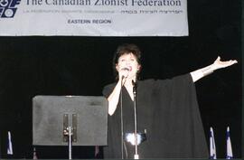 Interior snapshot of unidentified woman in front of music stands singing into microphone with outstretched arm. Banner reads "The Canadian Zionist Federation, Eastern Region."